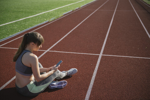 Sportlerin auf der Rennbahn sitzend mit Smartphone, lizenzfreies Stockfoto