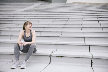 Junge Frau in Sportkleidung auf einer Betontribüne sitzend mit einer Flasche Wasser - AHSF00484