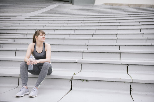 Young woman wearing sportswear sitting on concrete bleachers with a bottle of water - AHSF00484