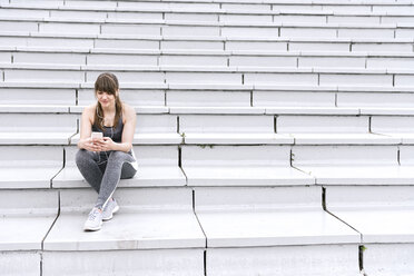 Junge Frau in Sportkleidung sitzt auf einer Betontribüne und hört Musik auf einem Smartphone - AHSF00485