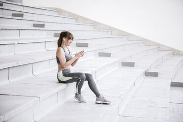 Junge Frau in Sportkleidung sitzt auf einer Betontribüne und hört Musik auf einem Smartphone - AHSF00489