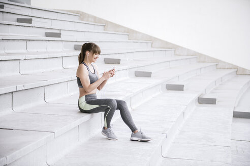 Young woman in sportswear sitting on concrete bleachers and listening music on a smartphone - AHSF00489