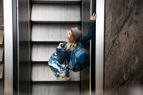 Woman with backpack and travelling bag standing on escalator looking up - HMEF00435