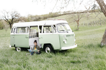 Young woman studying map, sitting in the grass, in front of her camper - HMEF00447