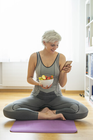 Frau, die zu Hause Yoga praktiziert, einen gesunden Obstsnack zu sich nimmt und ihr Smartphone benutzt, lizenzfreies Stockfoto