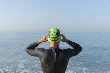 Triathlet bereitet sich auf das Schwimmen vor, setzt Badekappe und Schwimmbrille auf, Rückansicht - JPTF00115