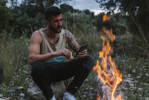 Mann sitzt am Lagerfeuer in ländlicher Landschaft und benutzt ein Mobiltelefon, lizenzfreies Stockfoto