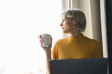 Woman looking out of window at home with laptop and coffee cup - FBAF00698