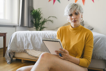 Portrait of woman sitting next to bed at home with notepad - FBAF00702