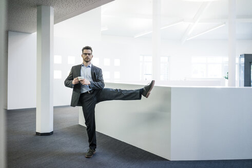 Geschäftsmann mit einer Tasse Kaffee auf einem Bein stehend im Büro - MOEF02177