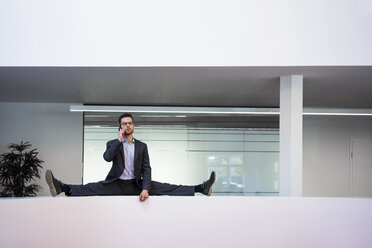 Businessman doing the splits on reception desk in office talking on cell phone - MOEF02182