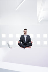 Businessman doing yoga in office next to laptop - MOEF02201