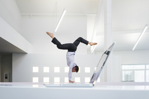 Businessman doing a handstand at laptop in office - MOEF02212