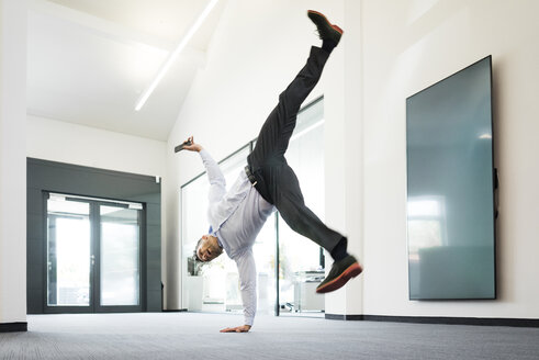 Businessman with cell phone doing a one-handed handstand on office floor - MOEF02214