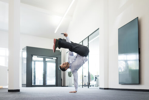 Geschäftsmann mit Mobiltelefon macht einen einhändigen Handstand auf dem Büroboden - MOEF02215