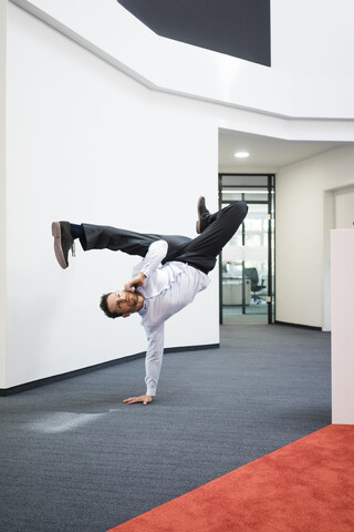 Geschäftsmann mit Mobiltelefon macht einen einhändigen Handstand auf dem Büroboden, lizenzfreies Stockfoto