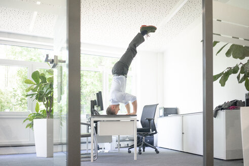 Geschäftsmann macht einen Kopfstand auf dem Schreibtisch im Büro - MOEF02231