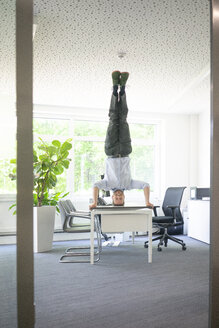 Businessman doing a headstand on desk in office - MOEF02232