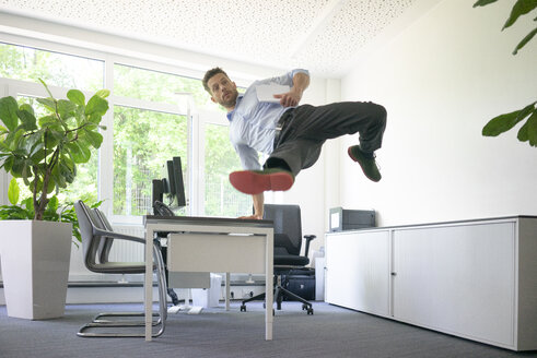 Geschäftsmann macht einen einhändigen Handstand auf dem Schreibtisch im Büro - MOEF02233