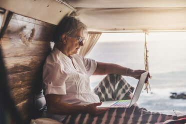 Senior woman sitting in vintage van by he sea, using laptop - SIPF02012