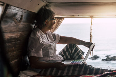 Senior woman sitting in vintage van by he sea, using laptop - SIPF02013