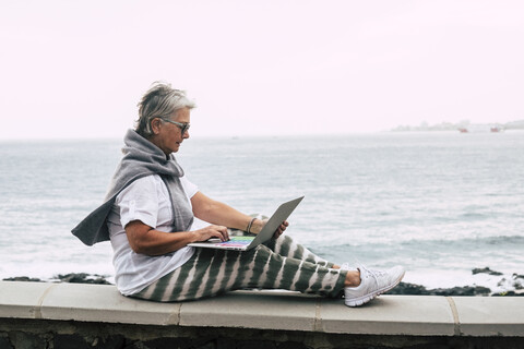 Ältere Frau sitzt auf einer Mauer am Meer und benutzt einen Laptop, lizenzfreies Stockfoto