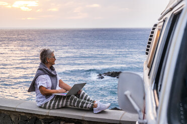 Senior woman travelling with a vintage van, sitting on wall by the sea, using laptop - SIPF02022