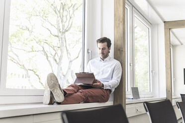 Junger Geschäftsmann sitzt auf der Fensterbank und benutzt ein digitales Tablet - UUF17700