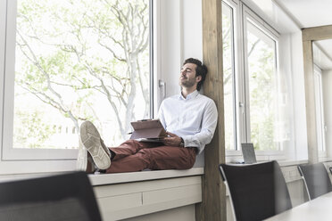 Junger Geschäftsmann mit digitalem Tablet, der auf der Fensterbank sitzt und eine Pause macht - UUF17702