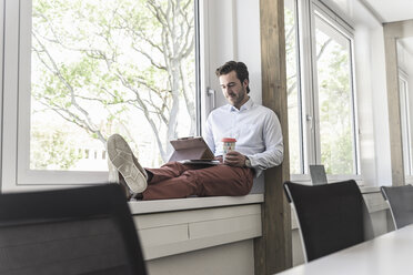 Young businessman sitting on windowsill, using digital tablet, drinking coffee - UUF17703