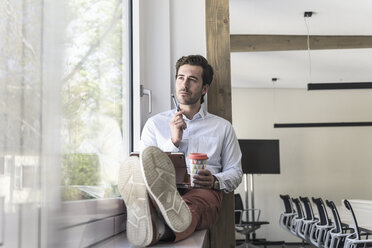 Junger Geschäftsmann sitzt auf der Fensterbank, benutzt ein digitales Tablet und trinkt Kaffee - UUF17704