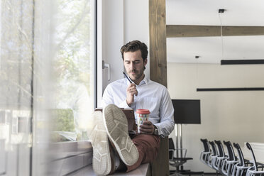 Young businessman sitting on windowsill, using digital tablet, drinking coffee - UUF17705