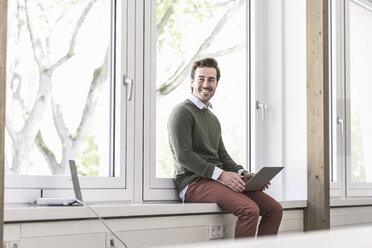 Young businessman sitting on windowsill, using laptop - UUF17711