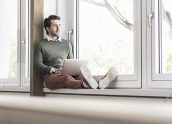 Young businessman sitting on windowsill, using laptop - UUF17712