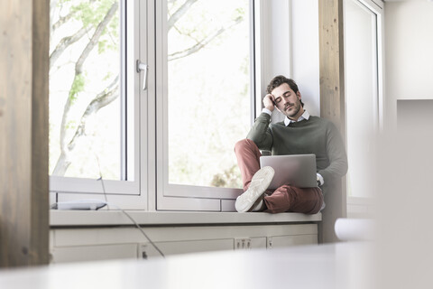 Young businessman with laptop sitting on windowsill, taking a break stock photo