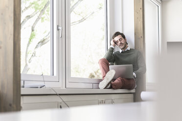 Junger Geschäftsmann mit Laptop, der auf der Fensterbank sitzt und eine Pause macht - UUF17713