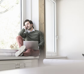 Young businessman with laptop sitting on windowsill, taking a break - UUF17714