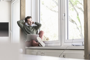 Young businessman sitting on windowsill, using laptop - UUF17715