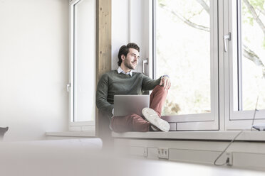 Young businessman sitting on windowsill, using laptop - UUF17716