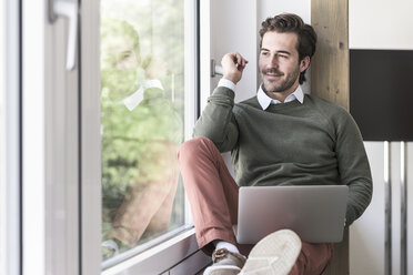 Young businessman sitting on windowsill, using laptop - UUF17717