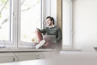 Young businessman with laptop sitting on windowsill, taking a break - UUF17718