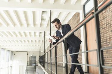 Young businessman standing on gallery in modern office building, using digital tablet - UUF17753
