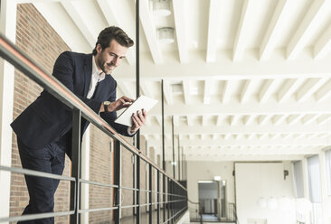 Young businessman standing on gallery in modern office building, using digital tablet - UUF17754