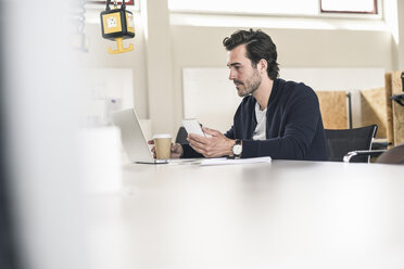 Young businessman sitting in office, using laptop and smartphone - UUF17790