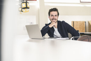 Entspannter junger Geschäftsmann mit Laptop, sitzend im Büro - UUF17794