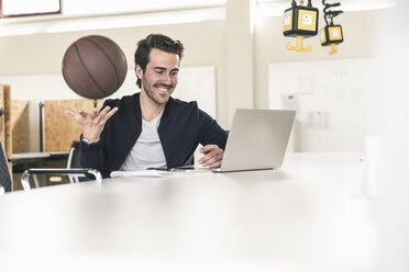 Young businessman using laptop, playing with a basketball - UUF17795