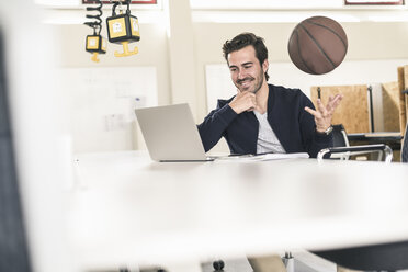 Young businessman using laptop, playing with a basketball - UUF17796