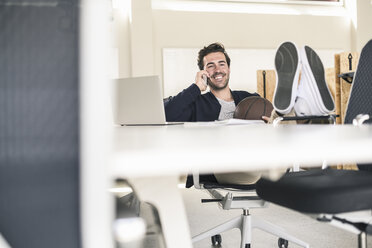 Young businessman sitting in office, with feet on desk, talking on the phone - UUF17797