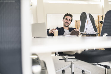 Junger Geschäftsmann sitzt im Büro, Füße auf dem Schreibtisch, trinkt Kaffee - UUF17798