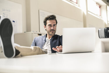 Young businessman working relaxed in modern office, using laptop with feet on desk - UUF17806